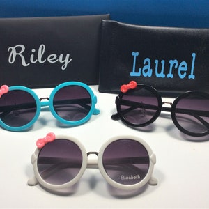 May include: Three pairs of round sunglasses with a pink bow on the side. The sunglasses are in blue, black, and white frames. The sunglasses are labeled with the names Riley, Laurel, and Elizabeth.