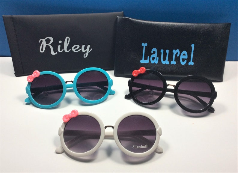 May include: Three pairs of round sunglasses with a pink bow on the side. The sunglasses are in blue, black, and white frames. The sunglasses are labeled with the names Riley, Laurel, and Elizabeth.