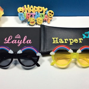 Puede incluir: Dos pares de gafas de sol arco&iacute;ris con monturas negras y amarillas. Las gafas de sol est&aacute;n en sus estuches, que son negros y tienen los nombres "Layla" y "Harper" escritos en ellos.