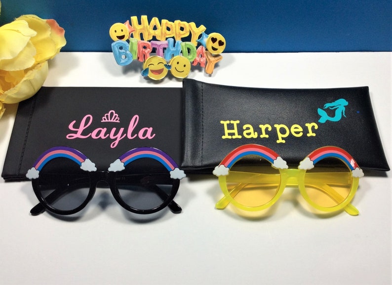 Puede incluir: Dos pares de gafas de sol arco&iacute;ris con monturas negras y amarillas. Las gafas de sol est&aacute;n en sus estuches, que son negros y tienen los nombres "Layla" y "Harper" escritos en ellos.