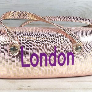May include: A rose gold, textured eyeglasses case with a handle and the word "London" in purple. The case has a metallic finish and a reptile-like pattern. The handle is attached with silver-coloured hardware.