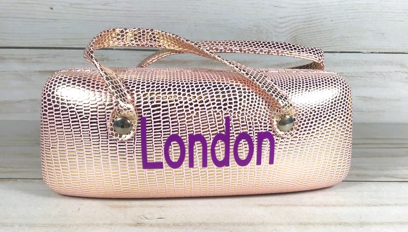 May include: A rose gold, textured eyeglasses case with a handle and the word "London" in purple. The case has a metallic finish and a reptile-like pattern. The handle is attached with silver-coloured hardware.