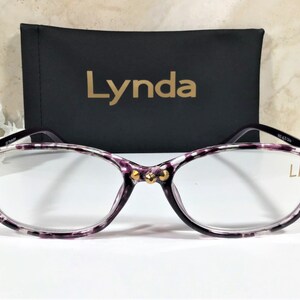 Puede incluir: Gafas con un patr&oacute;n morado y blanco, con detalles dorados en las monturas. Detr&aacute;s de las gafas hay un estuche negro con el nombre "Lynda" en letras doradas. Las lentes son transparentes.