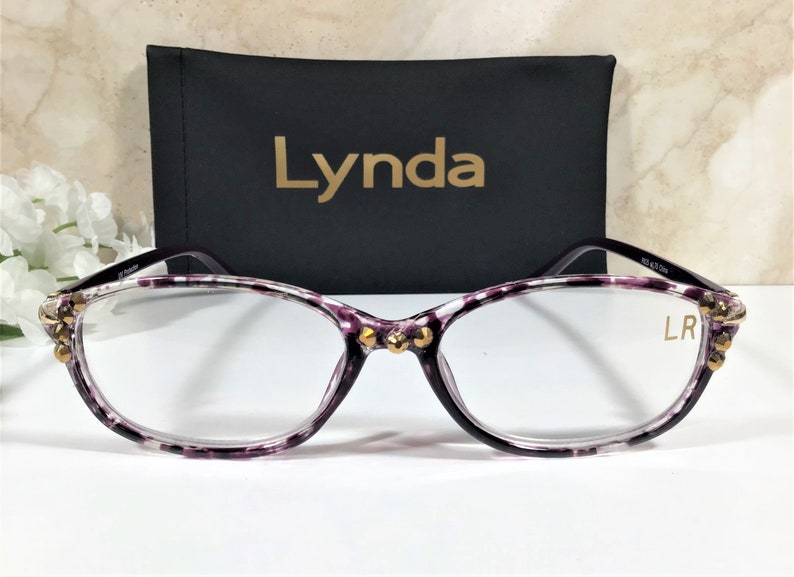 Puede incluir: Gafas con un patr&oacute;n morado y blanco, con detalles dorados en las monturas. Detr&aacute;s de las gafas hay un estuche negro con el nombre "Lynda" en letras doradas. Las lentes son transparentes.