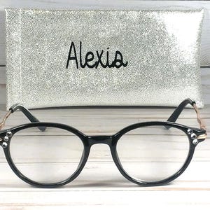 May include: Black-rimmed spectacles featuring clear lenses and small decorative rhinestones on the outer corners of the frames. The temples are metallic gold with a wavy design. Behind the glasses sits a silver glittery rectangular case with the name Alexia printed in black script.