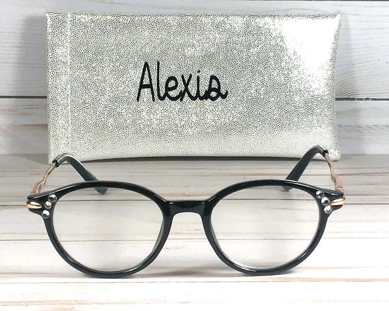 May include: Black-rimmed spectacles featuring clear lenses and small decorative rhinestones on the outer corners of the frames. The temples are metallic gold with a wavy design. Behind the glasses sits a silver glittery rectangular case with the name Alexia printed in black script.