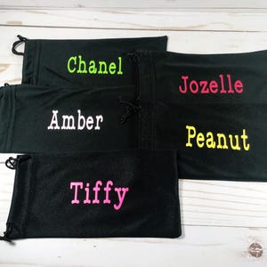 May include: Black drawstring pouches with embroidered names in various colors. The names include "Chanel" in green, "Jozelle" in pink, "Amber" in white, "Peanut" in yellow, and "Tiffy" in pink. The pouches are arranged on a light wood surface.