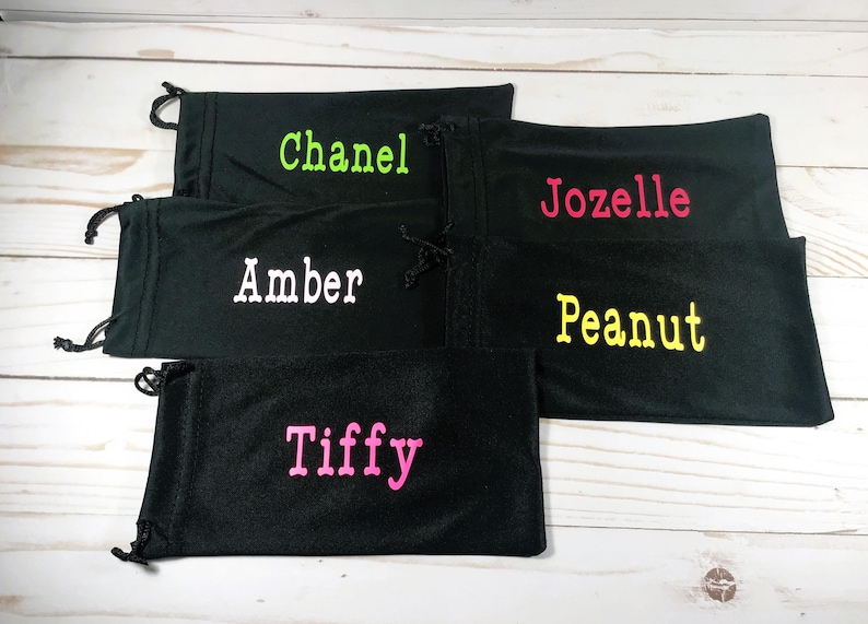 May include: Black drawstring pouches with embroidered names in various colors. The names include "Chanel" in green, "Jozelle" in pink, "Amber" in white, "Peanut" in yellow, and "Tiffy" in pink. The pouches are arranged on a light wood surface.