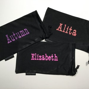 May include: Three black microfiber eyeglass cleaning pouches with different names embroidered on them in glitter: "Autumn" in purple, "Alita" in pink, and "Elizabeth" in pink.