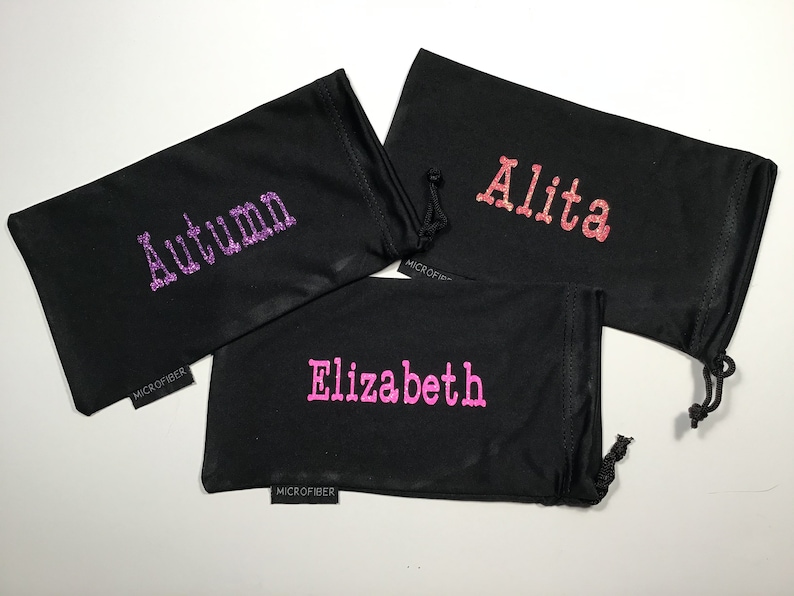 May include: Three black microfiber eyeglass cleaning pouches with different names embroidered on them in glitter: "Autumn" in purple, "Alita" in pink, and "Elizabeth" in pink.