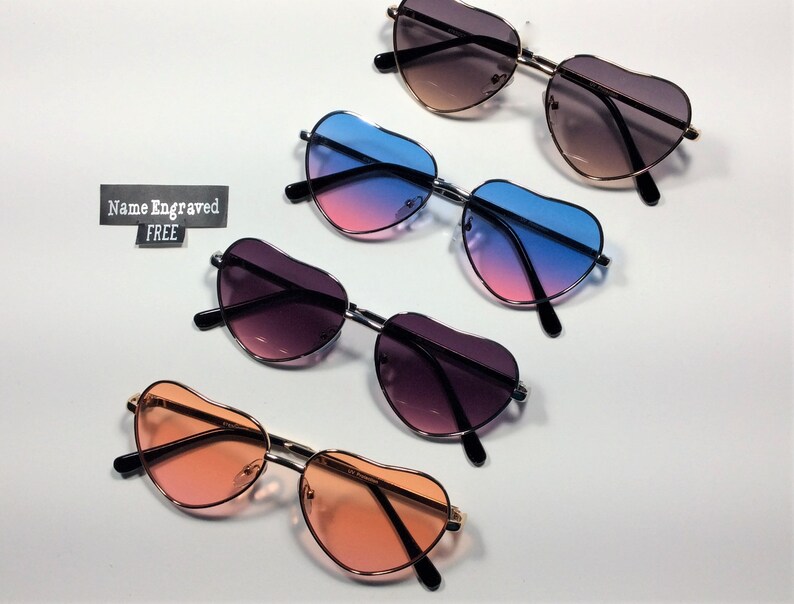 May include: Four pairs of heart-shaped sunglasses with different colored lenses. The lenses are pink and blue, pink and purple, brown and black, and orange. The text "Name Engraved FREE" is visible in the image.