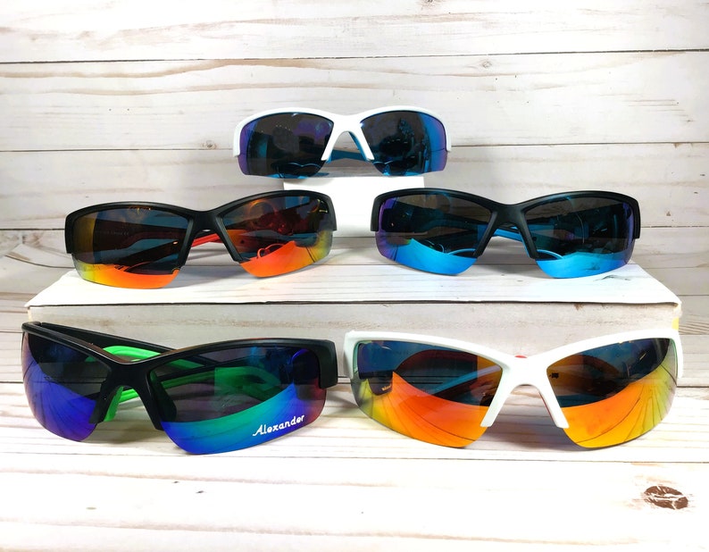 May include: Five pairs of athletic sunglasses with wrap-around frames and reflective lenses. The collection includes white and black frames with various lens colours, including blue, orange, and green. One pair features the word Alexander on the lens.