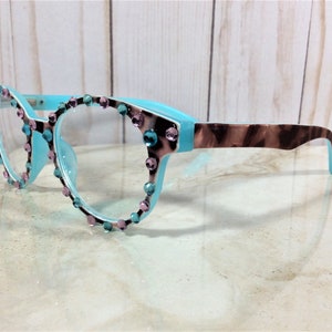 May include: A pair of eyeglasses with a light blue frame and clear lenses. The frame is adorned with pink and teal rhinestones around the lenses. The temples have a brown and tan tortoiseshell pattern.