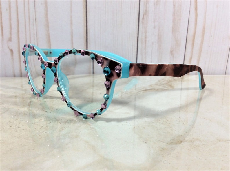May include: A pair of eyeglasses with a light blue frame and clear lenses. The frame is adorned with pink and teal rhinestones around the lenses. The temples have a brown and tan tortoiseshell pattern.