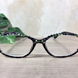 May include: Black-framed eyeglasses with clear lenses, adorned with green and dark blue rhinestones along the top rim. The glasses rest on a light wooden surface, with green foliage in the background.