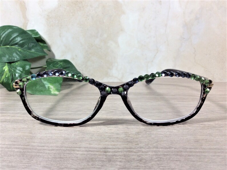 May include: Black-framed eyeglasses with clear lenses, adorned with green and dark blue rhinestones along the top rim. The glasses rest on a light wooden surface, with green foliage in the background.