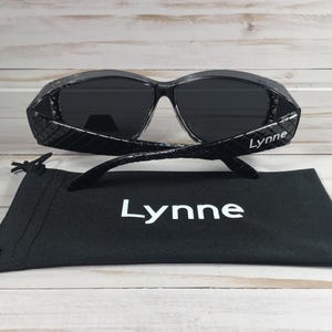 May include: Black sunglasses with a textured frame and dark lenses, resting on a black drawstring pouch. The sunglasses have the name "Lynne" printed on the temples and the pouch.
