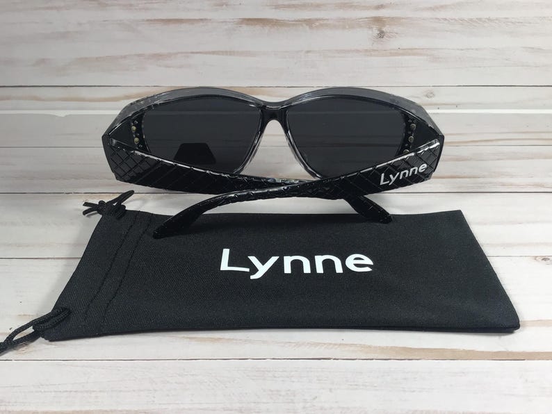 May include: Black sunglasses with a textured frame and dark lenses, resting on a black drawstring pouch. The sunglasses have the name "Lynne" printed on the temples and the pouch.