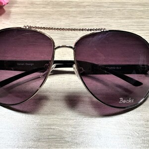 May include: Silver aviator sunglasses with dark purple lenses and rhinestones on the arms. The sunglasses have a chain detail connecting the frames. The text "Italian Design" and "Becks" are printed on the frames.