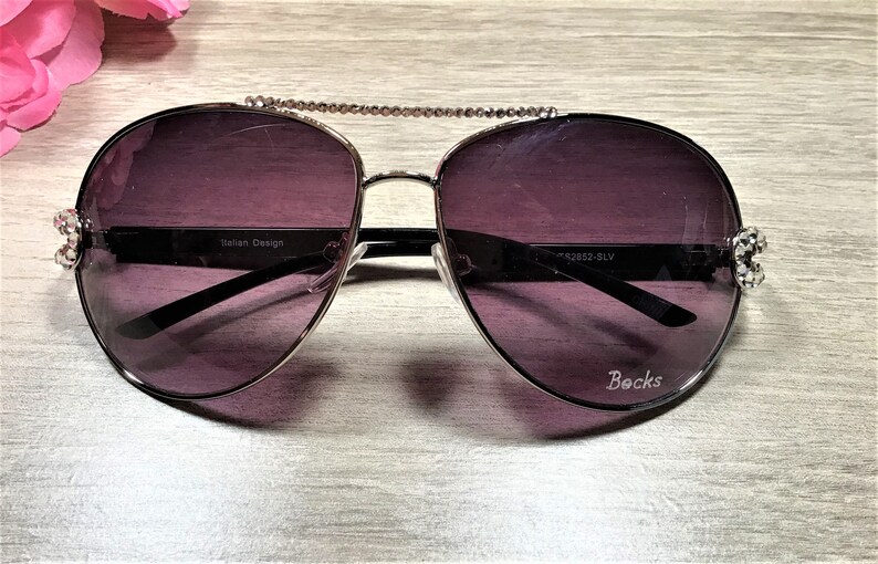 May include: Silver aviator sunglasses with dark purple lenses and rhinestones on the arms. The sunglasses have a chain detail connecting the frames. The text "Italian Design" and "Becks" are printed on the frames.