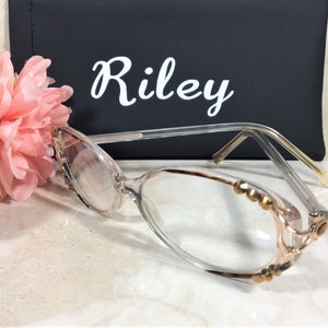 Puede incluir: Gafas con montura transparente y adornos dorados en los bordes. Las lentes son claras y las patillas son finas y transparentes. En el fondo, un estuche negro con el nombre "Riley" en escritura blanca.