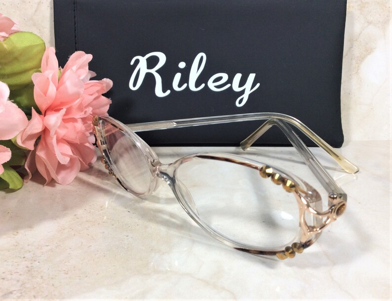 Puede incluir: Gafas con montura transparente y adornos dorados en los bordes. Las lentes son claras y las patillas son finas y transparentes. En el fondo, un estuche negro con el nombre "Riley" en escritura blanca.