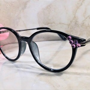 May include: Black eyeglasses with round frames, adorned with clusters of purple rhinestones at the temples. The glasses have clear lenses and silver hinges. The overall design is elegant and stylish.