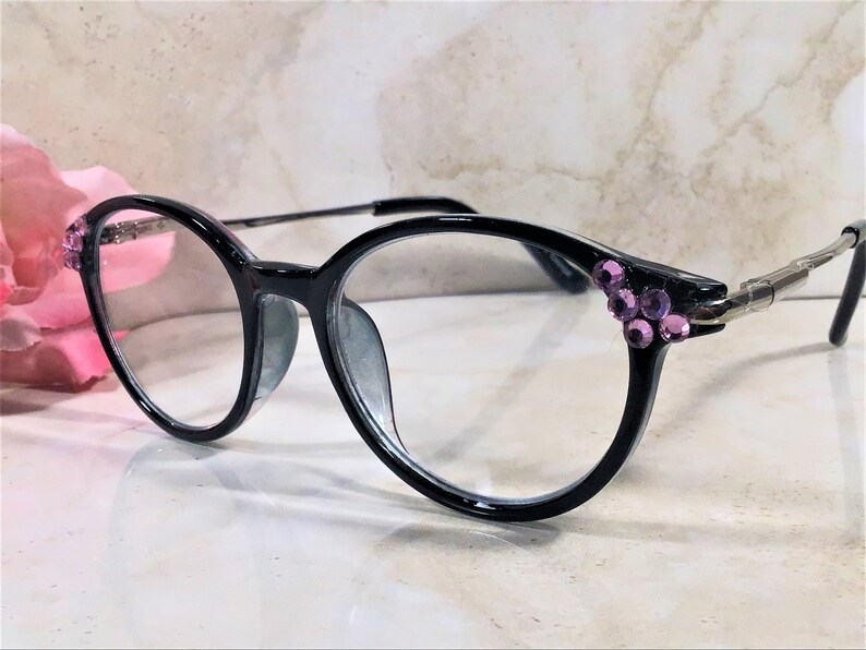 May include: Black eyeglasses with round frames, adorned with clusters of purple rhinestones at the temples. The glasses have clear lenses and silver hinges. The overall design is elegant and stylish.