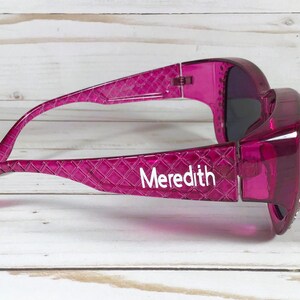May include: A pair of magenta sunglasses with a textured pattern on the arms. The name "Meredith" is printed in white on the left arm. The lenses are dark, and the frame is a transparent, glossy material.