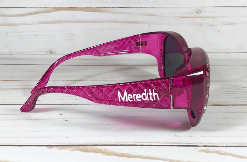 May include: A pair of magenta sunglasses with a textured pattern on the arms. The name "Meredith" is printed in white on the left arm. The lenses are dark, and the frame is a transparent, glossy material.