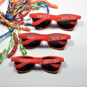 Puede incluir: Tres pares de gafas de sol de pl&aacute;stico rojo con lentes negros. Cada par tiene un nombre diferente impreso en la montura: Krista, Bowf y Taylan. Las gafas de sol tambi&eacute;n tienen el texto "COSTA 2021" impreso en la montura.