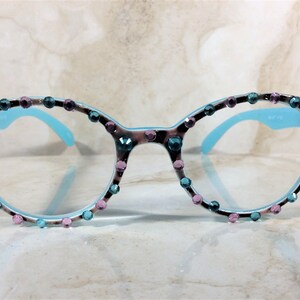 May include: Round-framed eyeglasses with a light blue interior and temples. The frames are decorated with a leopard print pattern and embellished with teal and pink rhinestones. The lenses are clear.