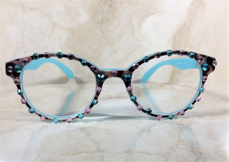 May include: Round-framed eyeglasses with a light blue interior and temples. The frames are decorated with a leopard print pattern and embellished with teal and pink rhinestones. The lenses are clear.