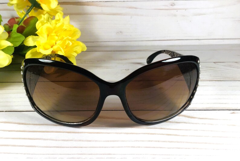 K&ouml;nnte beinhalten: Schwarze Cat-Eye-Sonnenbrille mit braunen Gl&auml;sern und Strasssteinen am Rahmen.