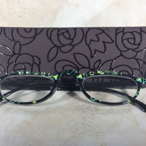 May include: Black-framed spectacles with a floral pattern and green gemstones along the top. The glasses are presented with a dark grey case decorated with a rose design.