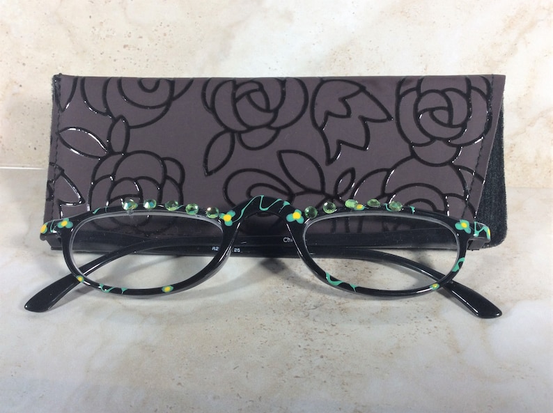 May include: Black-framed spectacles with a floral pattern and green gemstones along the top. The glasses are presented with a dark grey case decorated with a rose design.