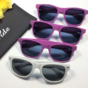 May include: Three pairs of sunglasses in a white background. The sunglasses are in three different colors: purple, pink, and white. The sunglasses have dark lenses. The sunglasses are in a black case with the word "Bride" written on it.