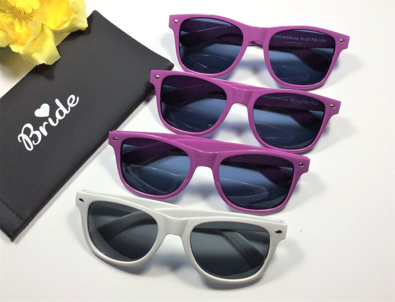 May include: Three pairs of sunglasses in a white background. The sunglasses are in three different colors: purple, pink, and white. The sunglasses have dark lenses. The sunglasses are in a black case with the word "Bride" written on it.