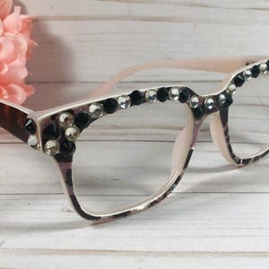 May include: A pair of eyeglasses with a light pink frame and clear lenses. The upper frame is decorated with black and clear rhinestones. The temples have a brown and white pattern. A pink flower is in the background.