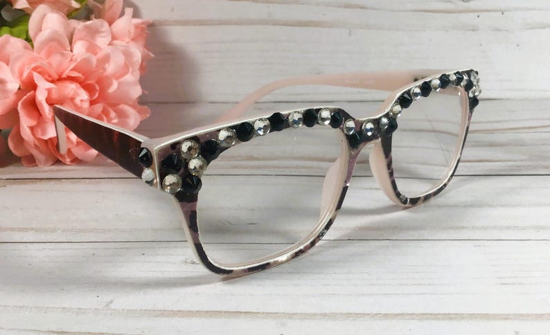 May include: A pair of eyeglasses with a light pink frame and clear lenses. The upper frame is decorated with black and clear rhinestones. The temples have a brown and white pattern. A pink flower is in the background.