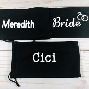 May include: Three black drawstring pouches with white text. One pouch reads "Meredith", another reads "Bride" with a ring graphic, and the third reads "Cici". The pouches are likely for gifts or wedding favors.