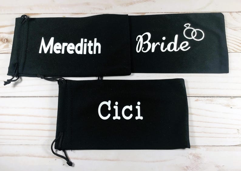 May include: Three black drawstring pouches with white text. One pouch reads "Meredith", another reads "Bride" with a ring graphic, and the third reads "Cici". The pouches are likely for gifts or wedding favors.