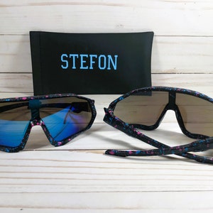 Puede incluir: Dos pares de gafas de sol negras con un dise&ntilde;o de pintura salpicada azul y rosa. Las gafas de sol tienen un estilo envolvente y est&aacute;n en una caja negra con la palabra "STEFON" impresa en azul.