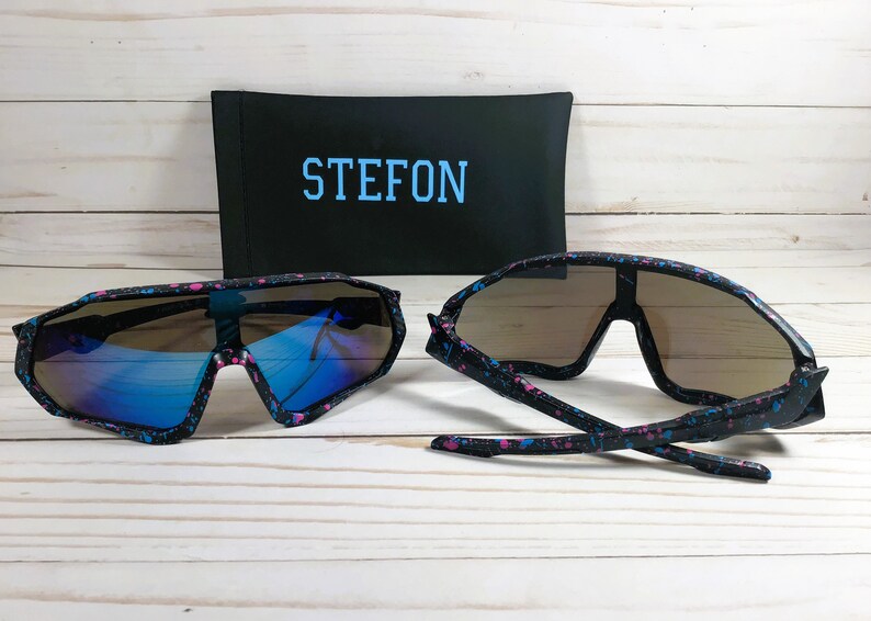 Puede incluir: Dos pares de gafas de sol negras con un dise&ntilde;o de pintura salpicada azul y rosa. Las gafas de sol tienen un estilo envolvente y est&aacute;n en una caja negra con la palabra "STEFON" impresa en azul.