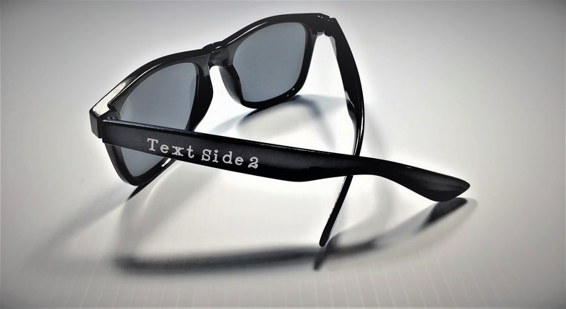 May include: Black sunglasses with a silver accent on the side. The sunglasses are folded in half and the text "Text Side 2" is engraved on the side.