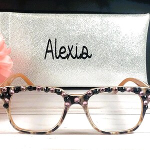 Op de afbeelding: Bril met een luipaardprint montuur en heldere glazen, versierd met zwarte en roze strass-steentjes. De naam "Alexis" staat in zwart schrift op een zilveren etui achter de bril. Links is een roze bloem.
