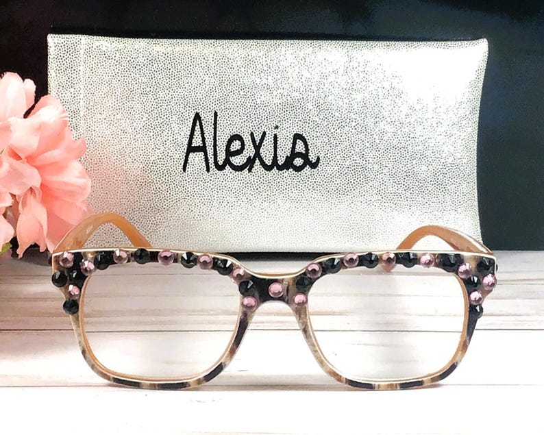 Op de afbeelding: Bril met een luipaardprint montuur en heldere glazen, versierd met zwarte en roze strass-steentjes. De naam "Alexis" staat in zwart schrift op een zilveren etui achter de bril. Links is een roze bloem.