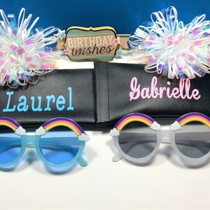 Puede incluir: Dos pares de gafas de sol arco&iacute;ris con detalles de nubes, una azul y una blanca, en estuches negros con los nombres "Laurel" y "Gabrielle" impresos en ellos. Los estuches est&aacute;n al lado de un cartel que dice "Birthday Wishes".