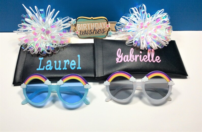 Puede incluir: Dos pares de gafas de sol arco&iacute;ris con detalles de nubes, una azul y una blanca, en estuches negros con los nombres "Laurel" y "Gabrielle" impresos en ellos. Los estuches est&aacute;n al lado de un cartel que dice "Birthday Wishes".