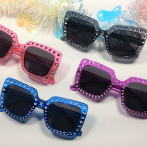 May include: Four pairs of square-shaped sunglasses with rhinestones around the frames. The sunglasses are in pink, purple, blue, and black.